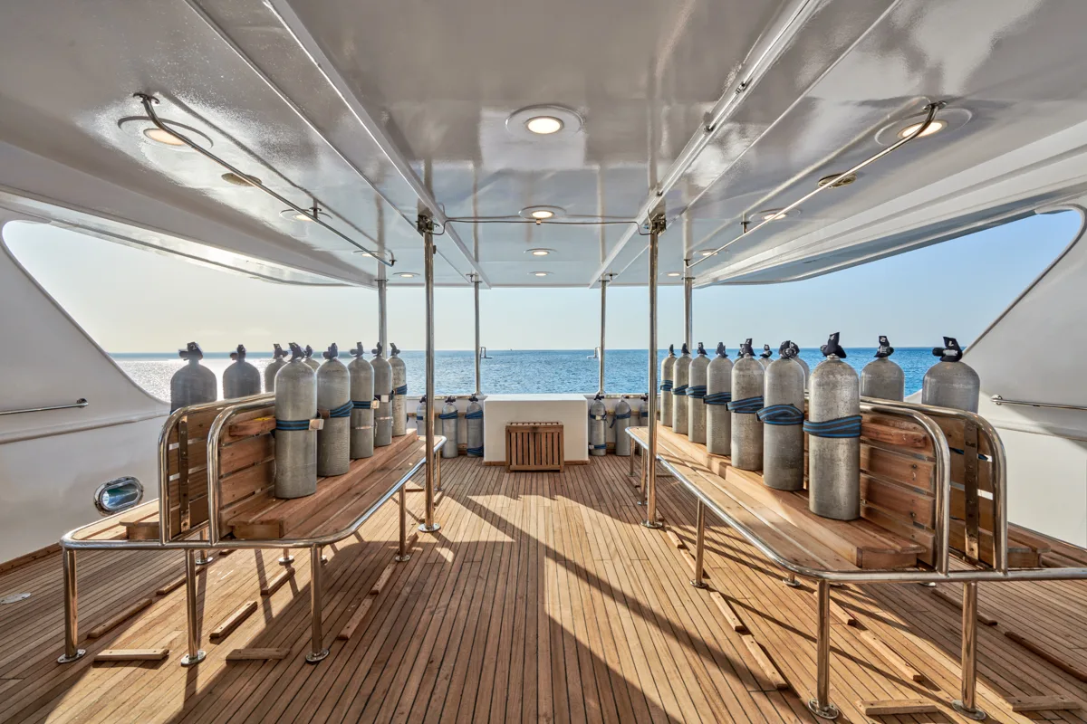 The covered dive deck of the liveaboard: dive cylinders strapped along teak benches on both sides, open to the sea at the stern